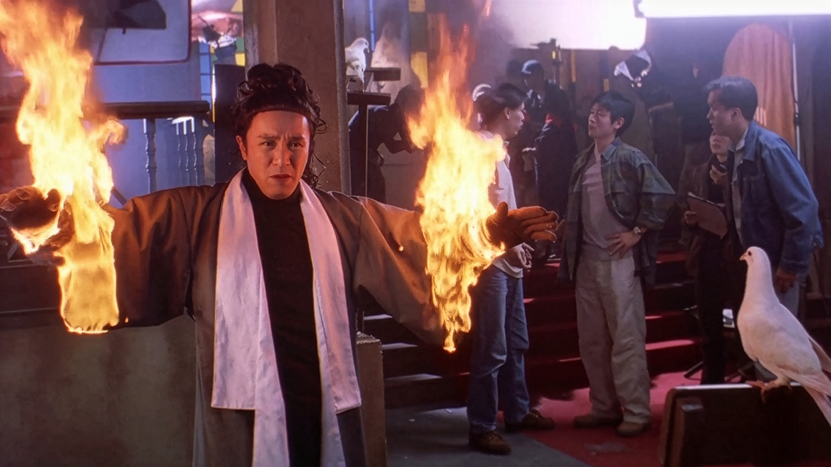 A scene from 'King of Comedy' where Stephen Chow, dressed in a historical costume, stands with an intense acting expression while his hands are engulfed in flames, as a film crew looks on nonchalantly in the background.