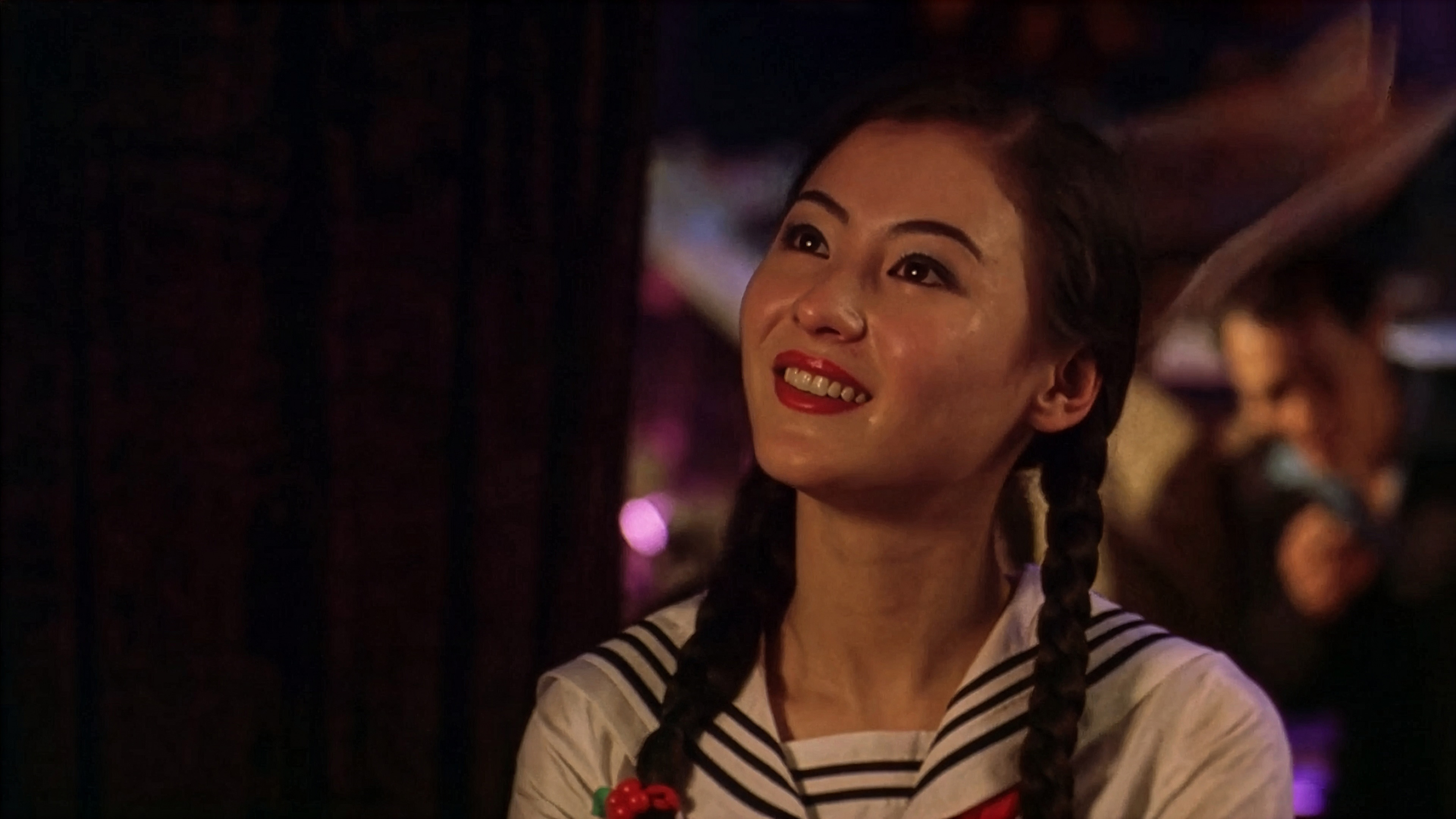 Upscaled still of a young Cecilia Cheung as the character Lau Piu-piu in 'King of Comedy.' She is dressed as a schoolgirl with two long braids and is looking upwards with a bright, hopeful smile in a dimly lit setting.